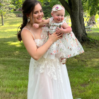 bride holding her Flower girl who's dressed in a white dress with embroidered flowers, a pink velvet headband with lace and silk roses accent and lace and silk rose shoe clips